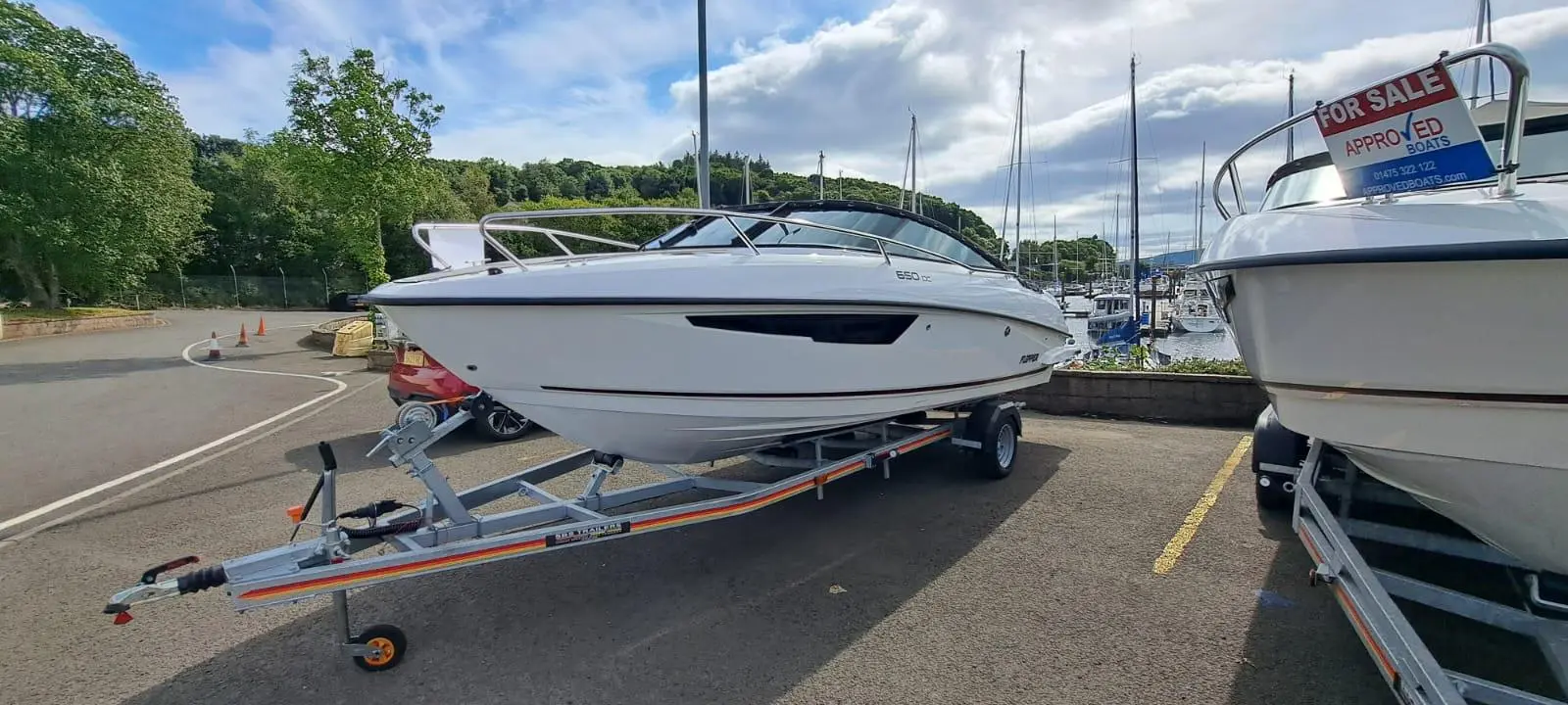 A white boat on a trailer in a marina, with "FOR SALE" sign and moored boats visible in the background.